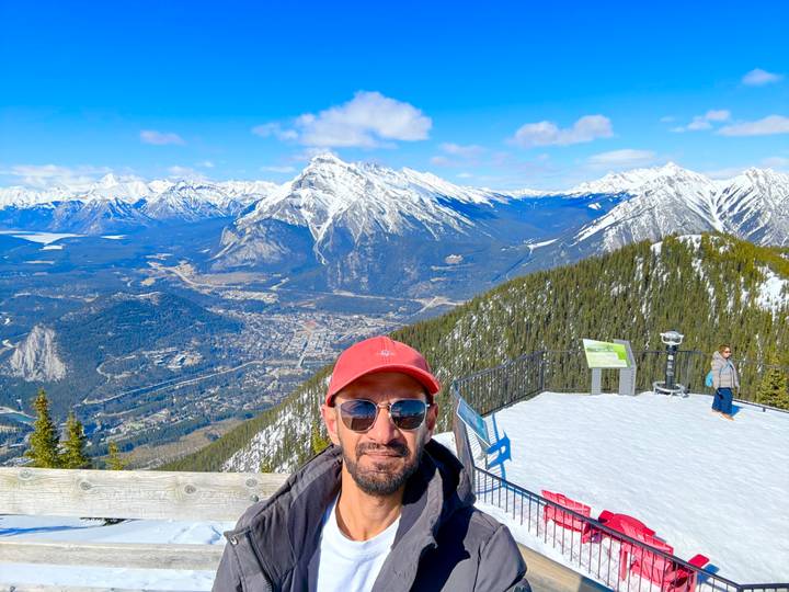 A man in a red cap poses on a snowy observation deck overlooking Banff townsite and dramatic snow-capped peaks.