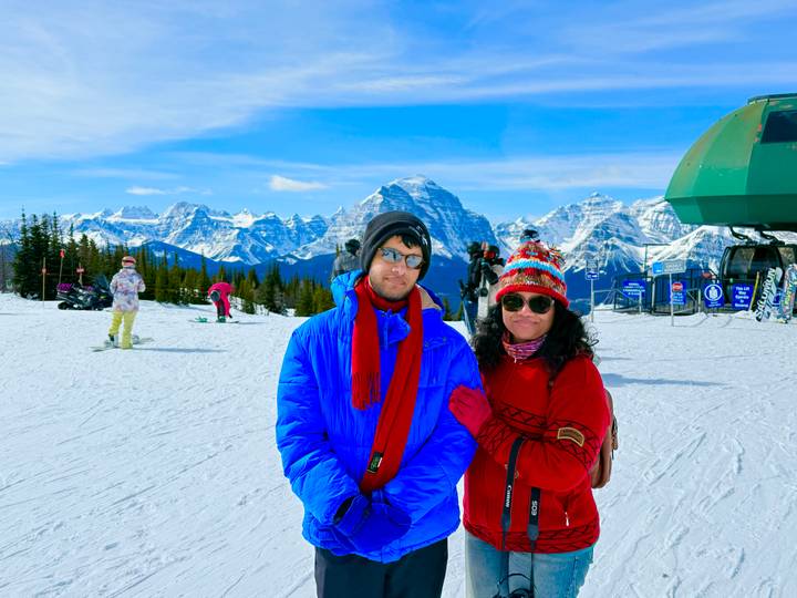 A young pair dressed in bright ski gear pose on a snowy ski field with impressive Rocky Mountain peaks in the background.