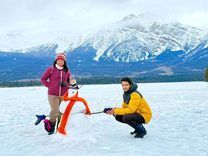 Two travelers build a snowman on a frozen lake with rugged snow-covered mountains towering in the distance.