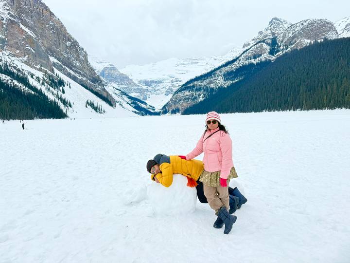 Two friends playfully pose on a vast snowy expanse framed by steep forested mountains and a distant glacier valley.
