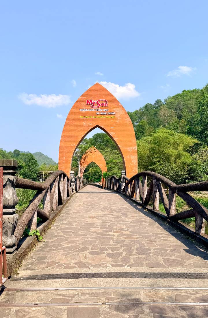 Stone footbridge leading to a tall terracotta archway framed by lush greenery and hills