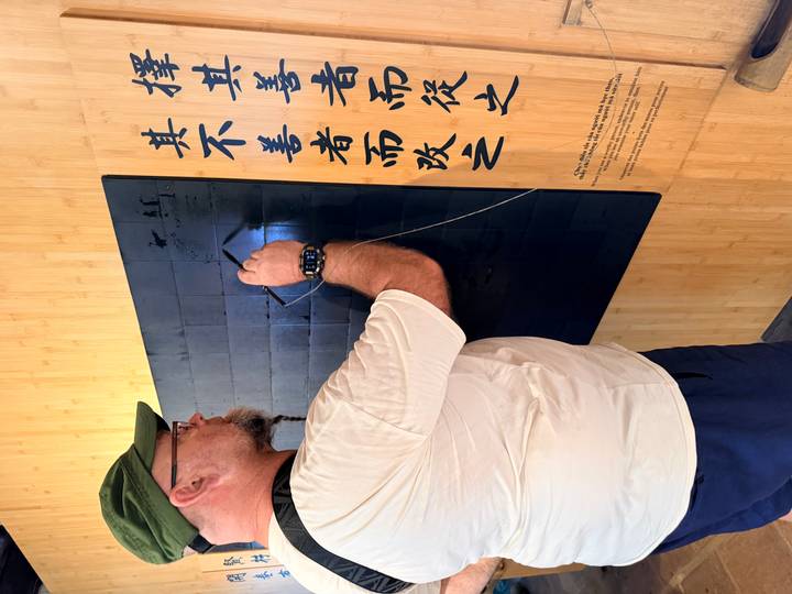 Man practicing calligraphy on a black tile board inside a cultural exhibit