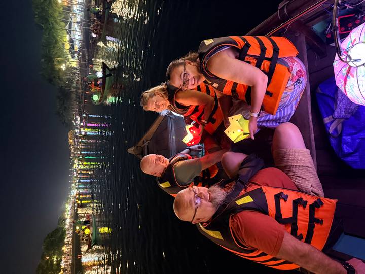 Four tourists in life vests seated in a wooden boat releasing paper lanterns onto a river at night