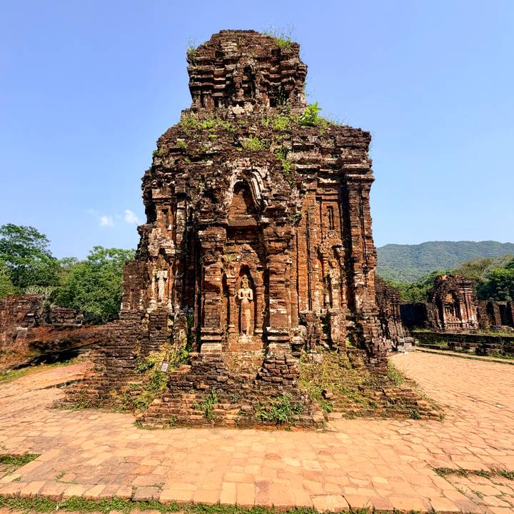 Ancient red-brick Hindu temple ruins surrounded by greenery and distant mountains under clear skies