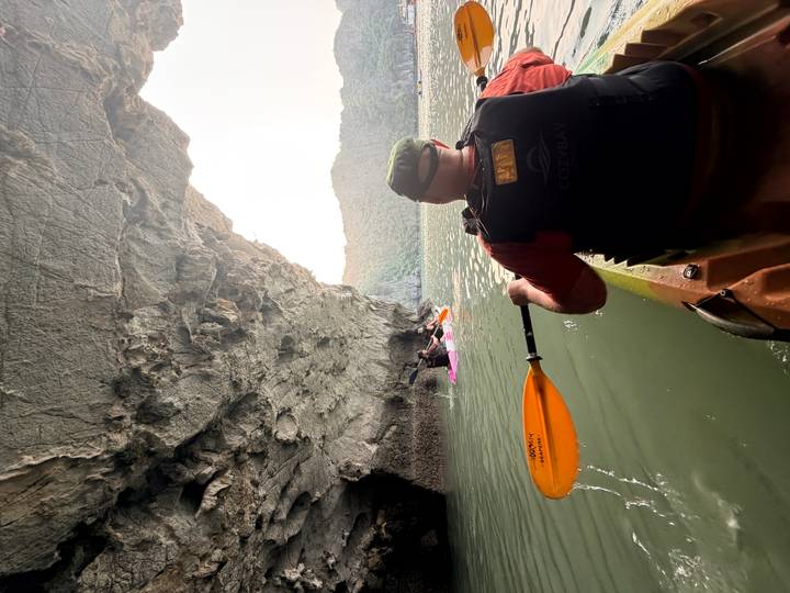 Kayaker paddling from inside a limestone cave toward open bay waters with cliffs beyond