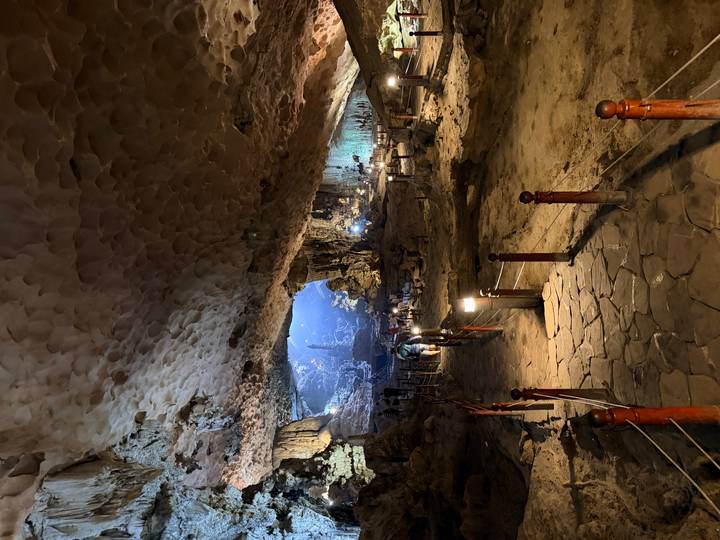Illuminated walkway descending into a vast limestone cave chamber with visitors in the distance