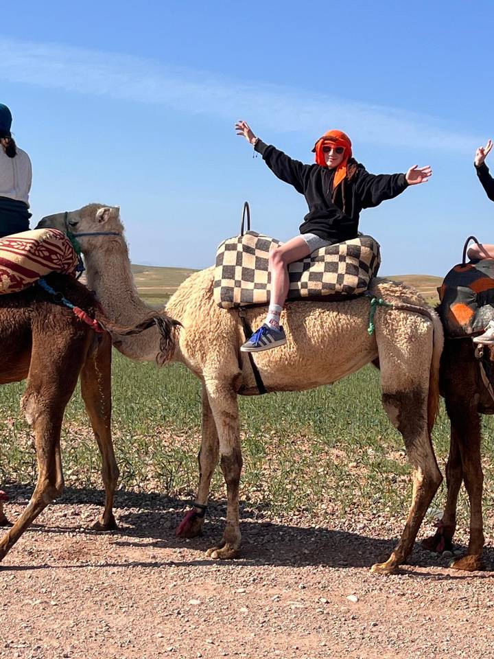 Young rider seated on a saddled camel in an open green field with gentle hills in the distance.