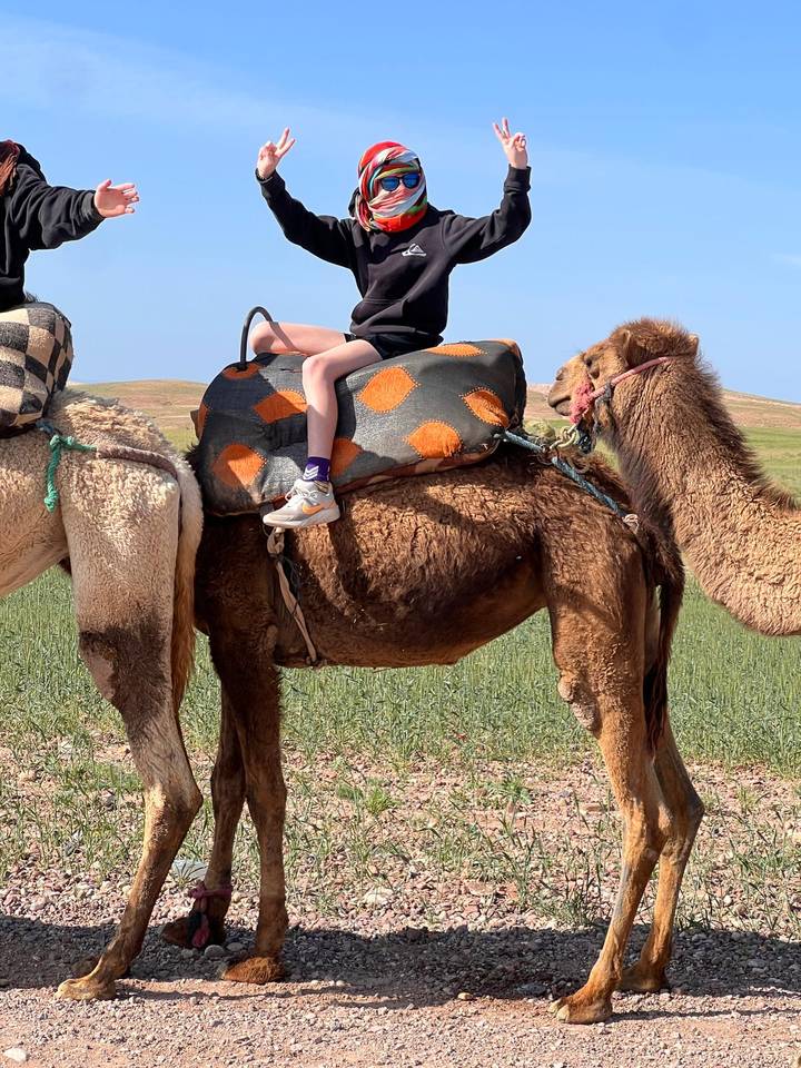 Close-up of a child sitting on a camel with patchwork saddle blankets in a grassy desert plain.