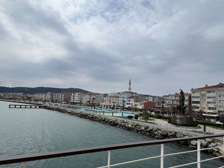 Seaside Turkish town with mosque, waterfront promenade and distant wooded hills under gray skies.