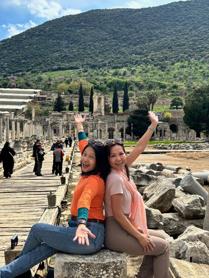 Two excited women posing with arms raised amid the ancient marble ruins of Ephesus.