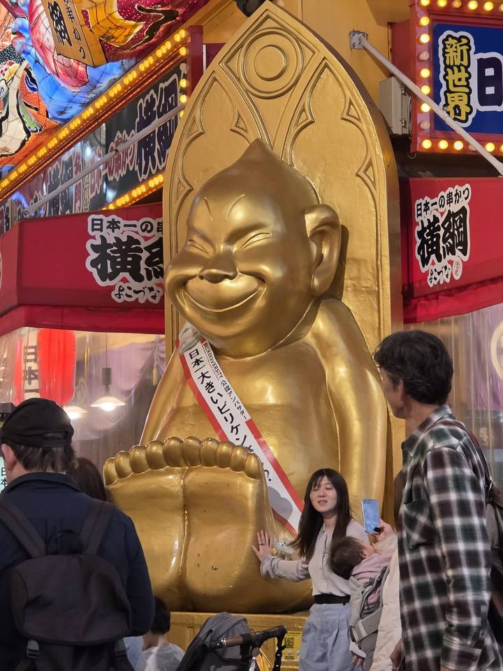 Golden Billiken statue displayed in a bustling Japanese street market while onlookers pass by.