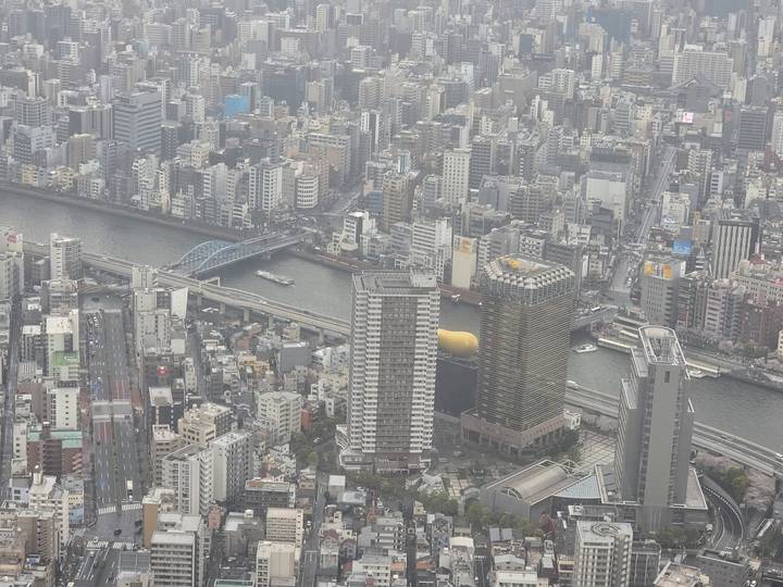 Aerial cityscape view over dense Tokyo buildings and the Sumida River with bridges and boats.