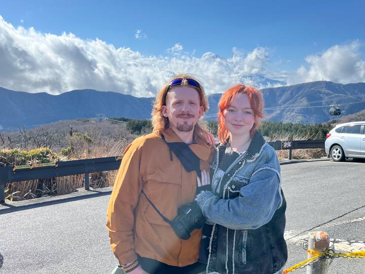 Young couple posing on mountain viewpoint with clouds, cable car and parked car behind.
