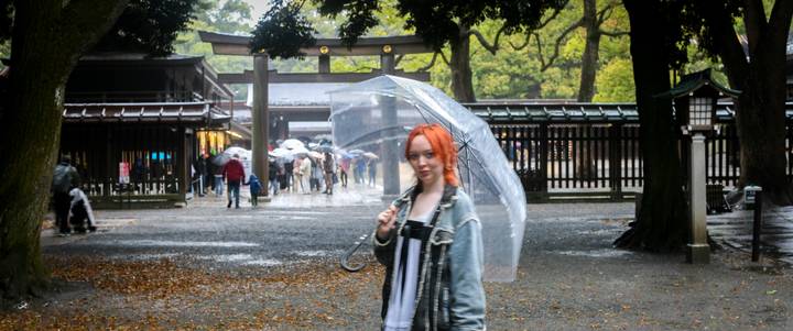 Young woman with transparent umbrella walking through rainy shrine courtyard.