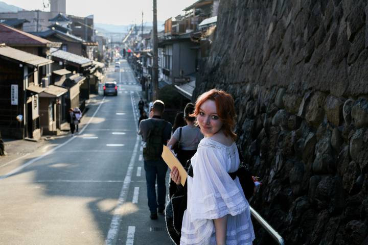 Red-haired traveler in white dress walking down traditional street toward distant town centre.
