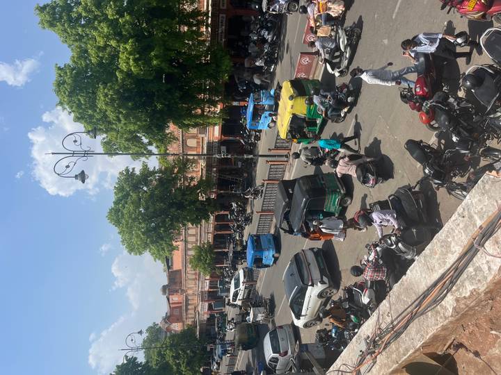 Busy Jaipur street scene with tuk-tuks, motorbikes and shoppers beneath pink sandstone facades.