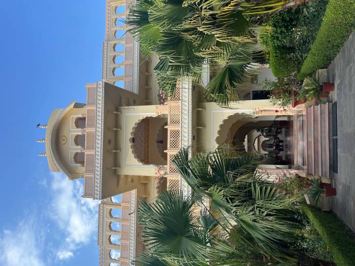 Grand heritage haveli with layered balconies framed by lush palm trees under clear sky.
