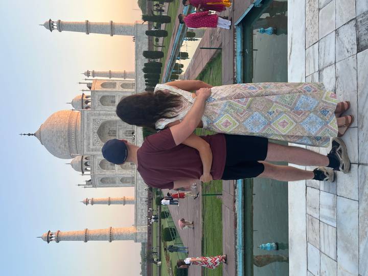 Couple stands arm-in-arm facing the Taj Mahal at sunset from the marble plinth.