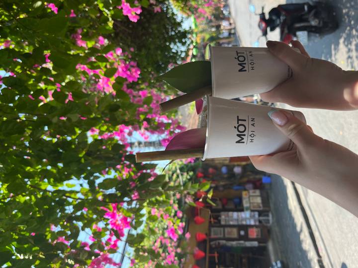 Two hands toast with signature herbal drinks in branded cups beneath bright bougainvillea on a sunny Hoi An street.