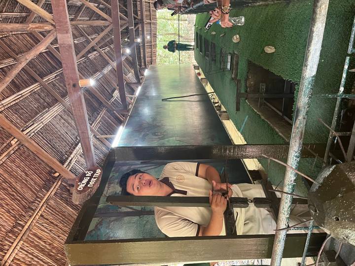 Guide demonstrating a Viet Cong door trap at the Cu Chi tunnels exhibit inside a thatched shelter.