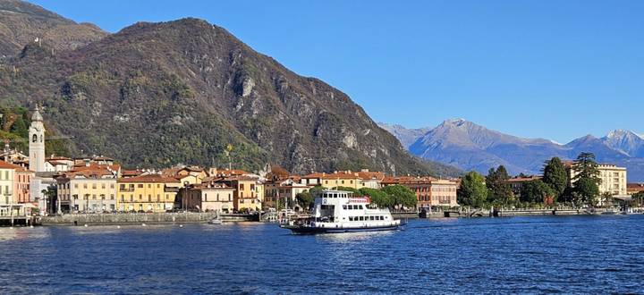Ferry boat gliding across the deep blue waters of a lake bordered by colorful Italian lakeside houses and steep wooded mountains.