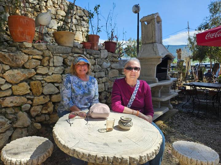 Two mature women relaxing at an outdoor stone-walled café table under potted plants and blue sky.