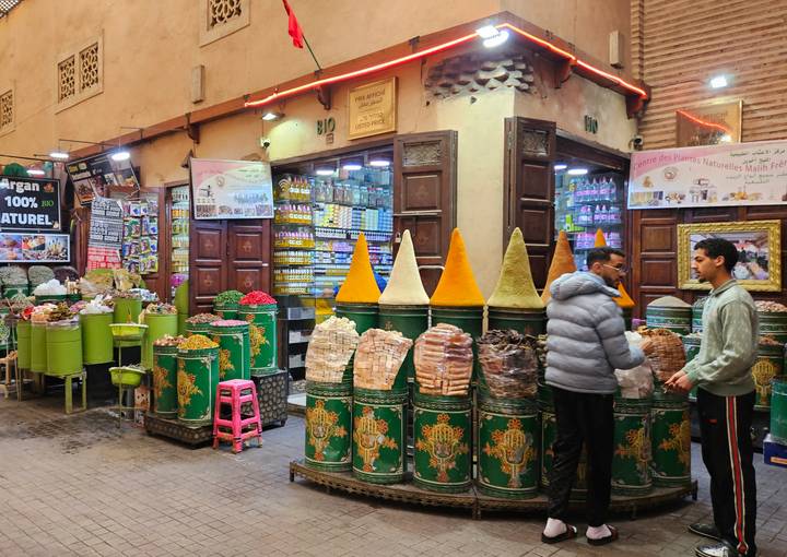 Spice and sweets stall in a Moroccan market lined with tall green barrels and colorful cones.