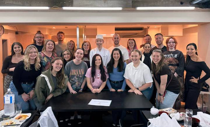 Large tour group poses with a sushi chef after a cooking class in a bright studio.