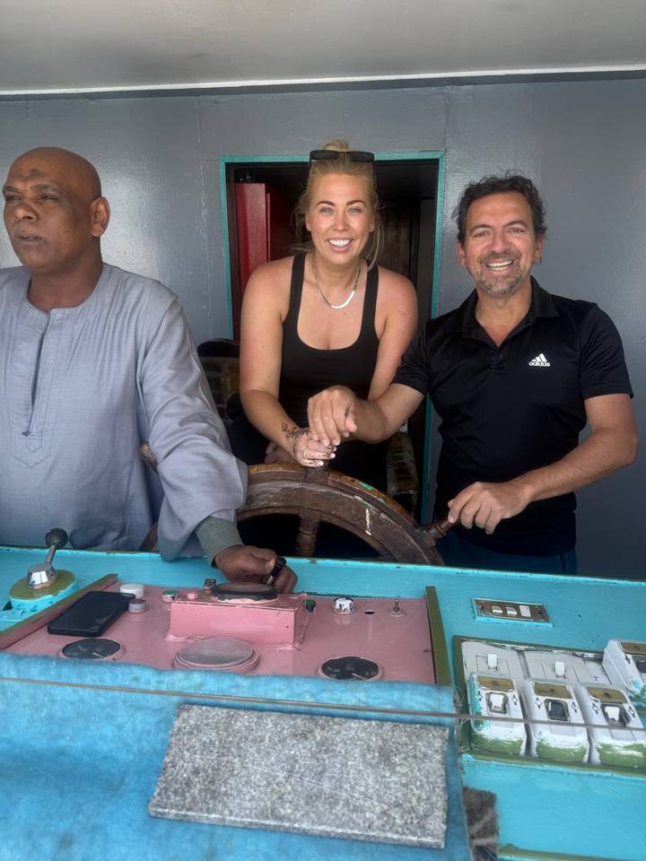 Smiling tourists with a local captain stand at the wooden helm of a Nile boat.