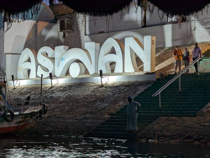 Illuminated ‘ASWAN’ sign on riverside steps at night with tourists posing nearby.