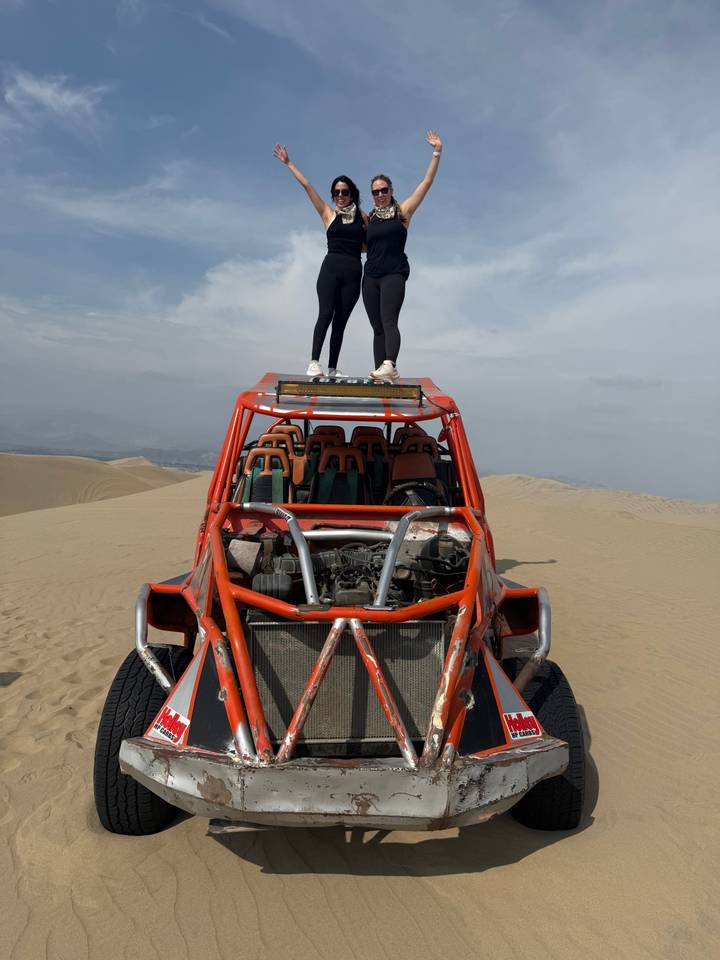 Two people stand on the roof rack of an orange dune buggy amidst vast desert sand dunes.