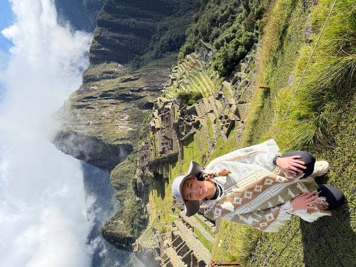 Traveller kneels smiling with the iconic ruins of Machu Picchu and Huayna Picchu peak shrouded in mist behind.