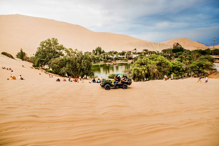 Dune buggy rides down golden sand dunes toward the lush oasis lagoon of Huacachina with scattered visitors relaxing.