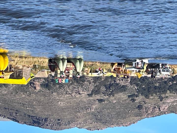 Floating reed island community with colourful UROS sign and tour boats on bright blue Lake Titicaca.