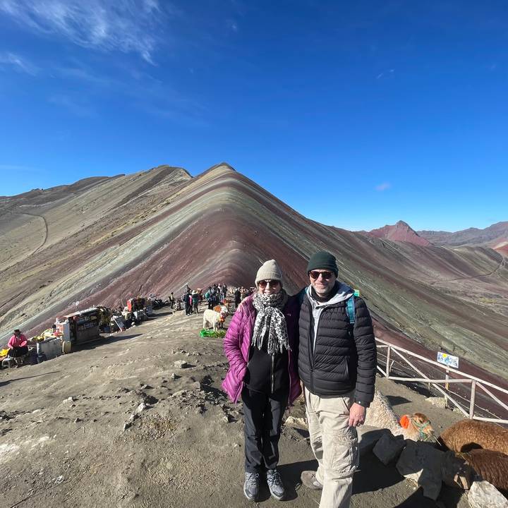 Two bundled travellers pose in front of the colourful striped ridge of Rainbow Mountain under clear skies.