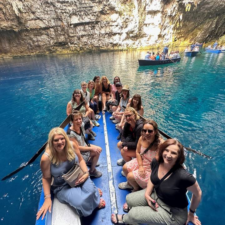 Group of women seated in a narrow rowboat on dazzling turquoise water inside a sunlit cave or sinkhole.