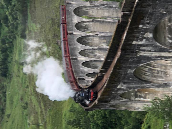 Steam train crossing a tall stone viaduct through green hills with smoke trailing.