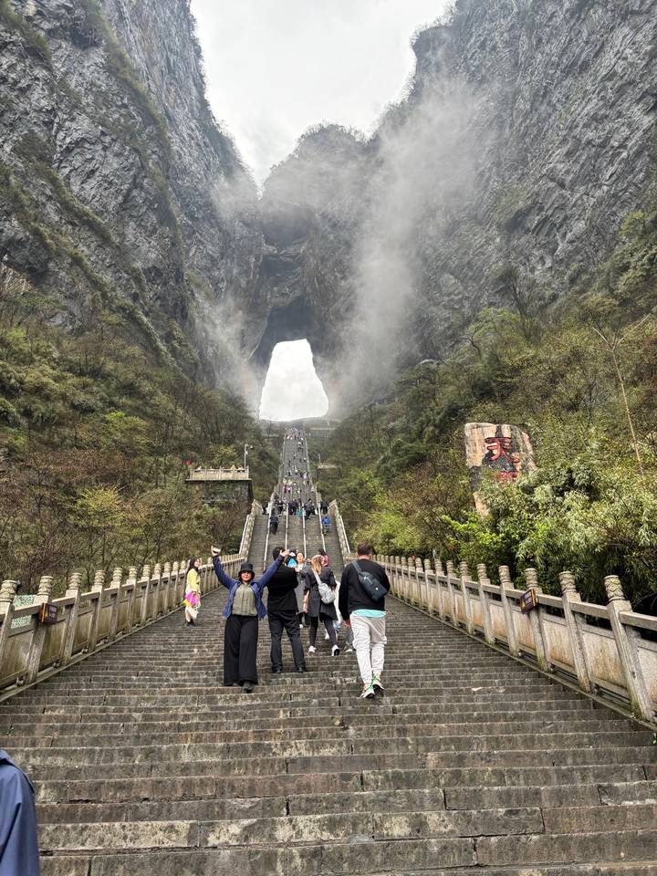 Grand stone staircase leads up to Tianmen Cave surrounded by misty cliffs while visitors climb and pose for photos.