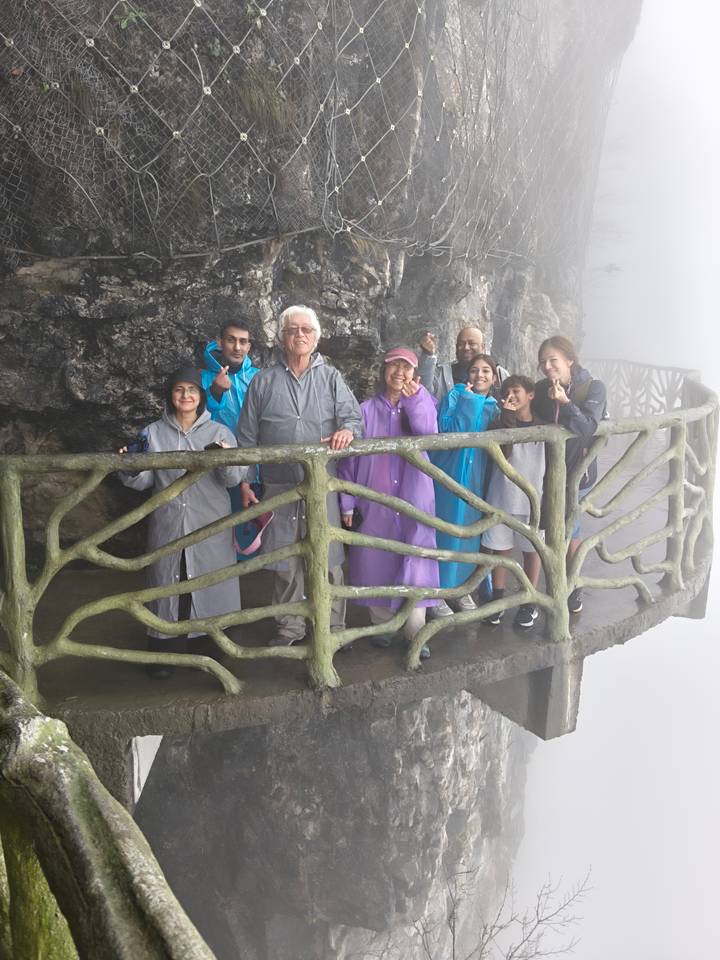 Smiling multi-generational group in colorful rain ponchos posing on a carved stone balcony in the mist.