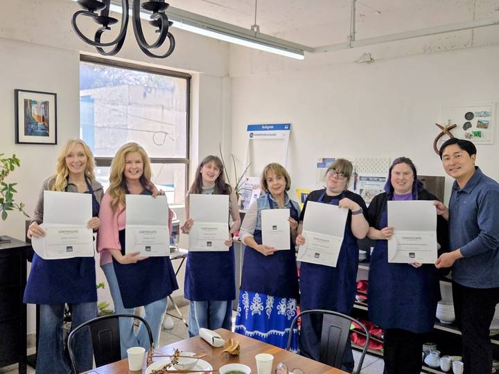 Seven participants wearing aprons proudly hold certificates inside a bright classroom after a cultural workshop.