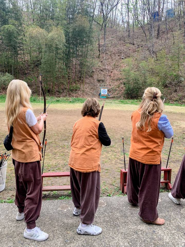 Three women in matching vests practice archery outdoors, aiming their bows toward a target on a forest range.