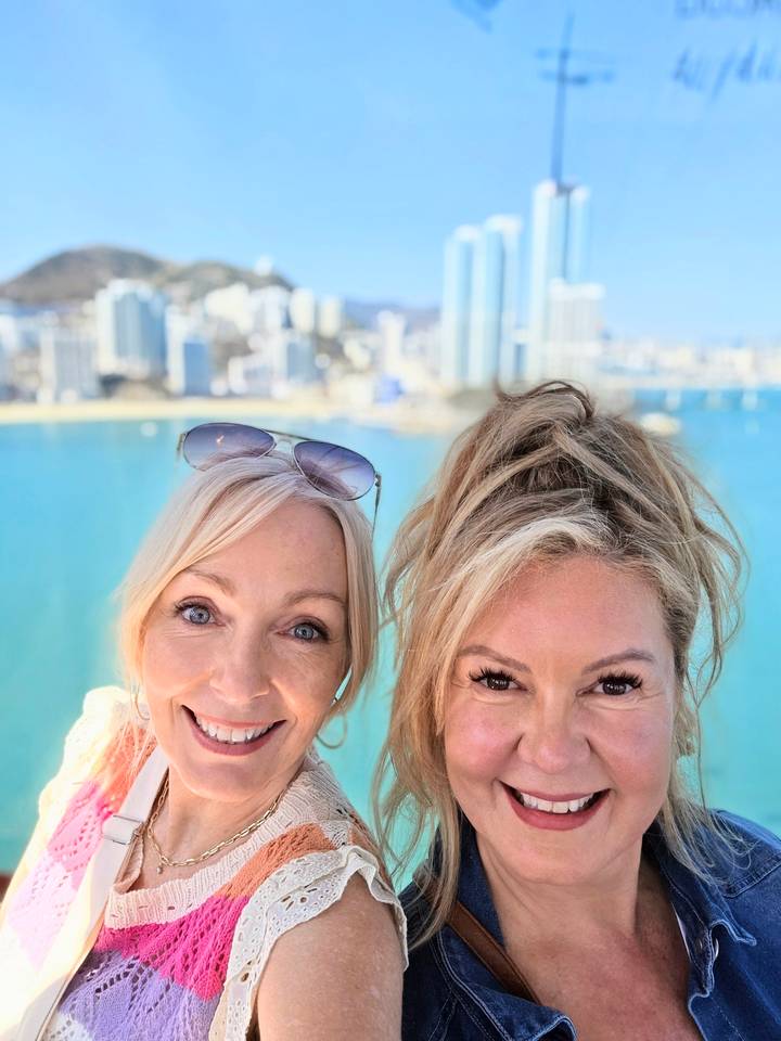 Close-up selfie of two smiling women with turquoise bay water and Busan city skyline blurred in the background.
