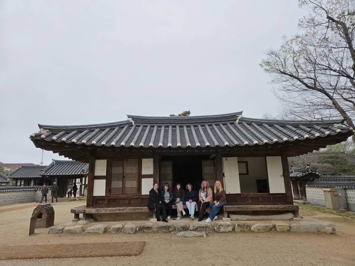 Travelers sit on the porch of a traditional Korean hanok with tiled roof inside a heritage village.