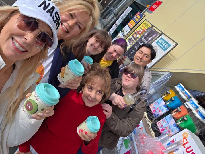 A cheerful group takes a selfie holding takeaway drinks outside a bustling Korean street stall.