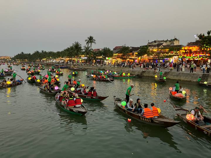 Dozens of small boats with colorful lanterns and life-jacketed passengers celebrating on a riverside at dusk.