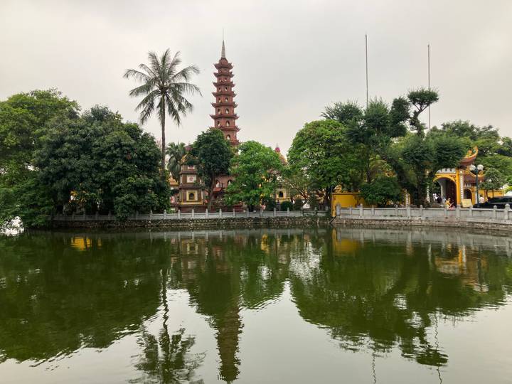Tall red-brick Tran Quoc Pagoda and lush greenery reflected in a small lake on a cloudy day.
