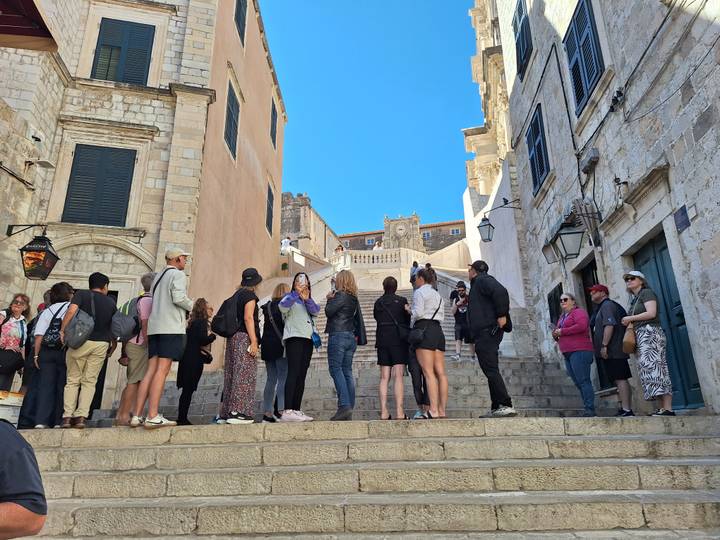 Tourists queue on stone steps between historic limestone buildings under a clear sky.