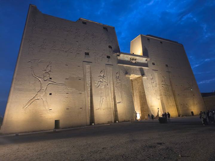 Illuminated pylons of the ancient Temple of Edfu stand against the deep blue twilight sky.