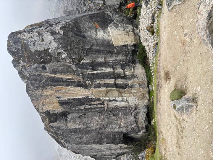 Massive granite boulder with streaked mineral patterns standing beside a rough mountain trail on a cloudy day.