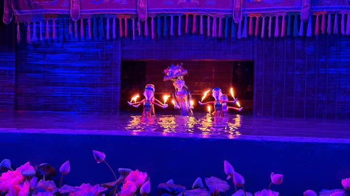 Colorful water-puppet performance with three puppets holding torches on a small watery stage under blue theatrical lighting.